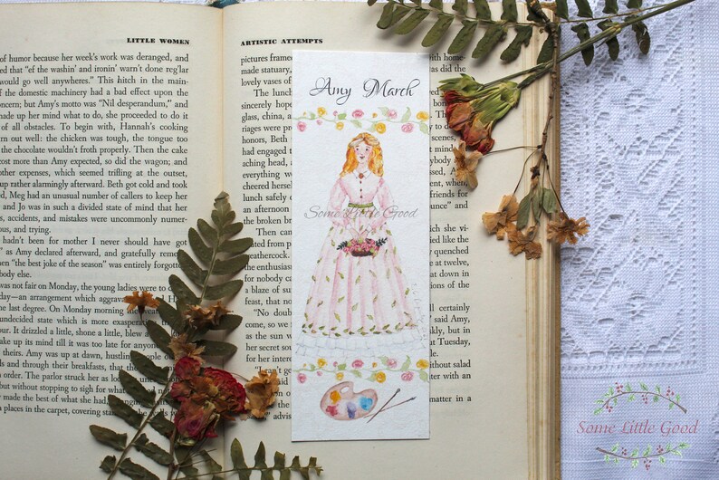 May include: A watercolor bookmark featuring a girl in a pink dress holding a bouquet of flowers. The bookmark has the text "Amy March" and "Some Little Good" on it. The background is a light blue with a watercolor painting of a palette and paintbrushes.