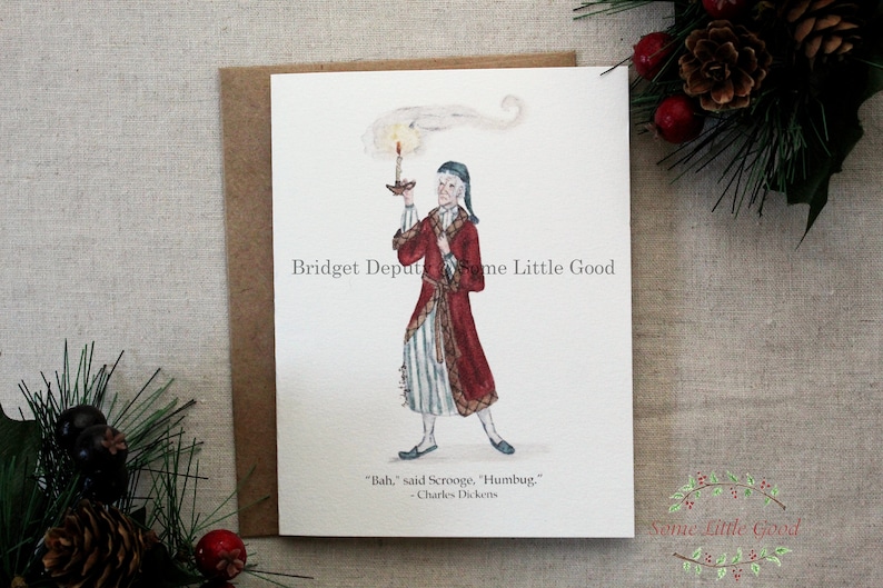 Pu&ograve; includere: Un biglietto di Natale con un'illustrazione ad acquerello di Ebenezer Scrooge che tiene in mano una candela, indossa un cappotto rosso e una cuffia da notte. La citazione "Bah", disse Scrooge, "Humbug", &egrave; sotto l'immagine. Il biglietto &egrave; su una busta marrone.