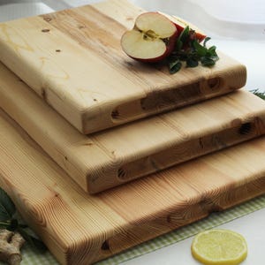 May include: A stack of three wooden cutting boards with rounded edges. The boards are made of light-colored wood and have a natural grain pattern. A sliced apple and sprigs of herbs rest on top. The boards are on a green and white checkered surface.