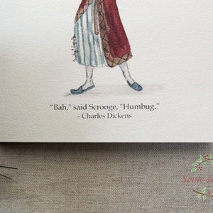 Pu&ograve; includere: Illustrazione ad acquerello di una figura in costume d'epoca, con un mantello rosso e pantaloni a righe. La citazione "Bah," disse Scrooge, "Humbug." - Charles Dickens &egrave; stampata sotto l'immagine su una carta bianca. &Egrave; visibile una busta marrone.