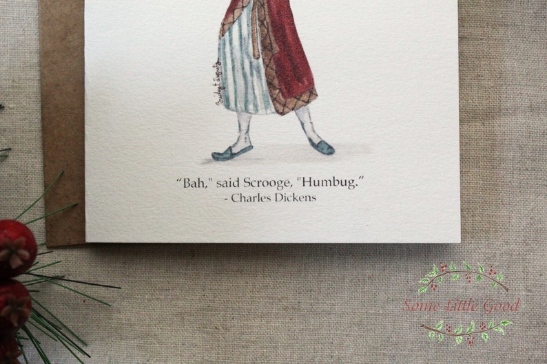 Pu&ograve; includere: Illustrazione ad acquerello di una figura in costume d'epoca, con un mantello rosso e pantaloni a righe. La citazione "Bah," disse Scrooge, "Humbug." - Charles Dickens &egrave; stampata sotto l'immagine su una carta bianca. &Egrave; visibile una busta marrone.
