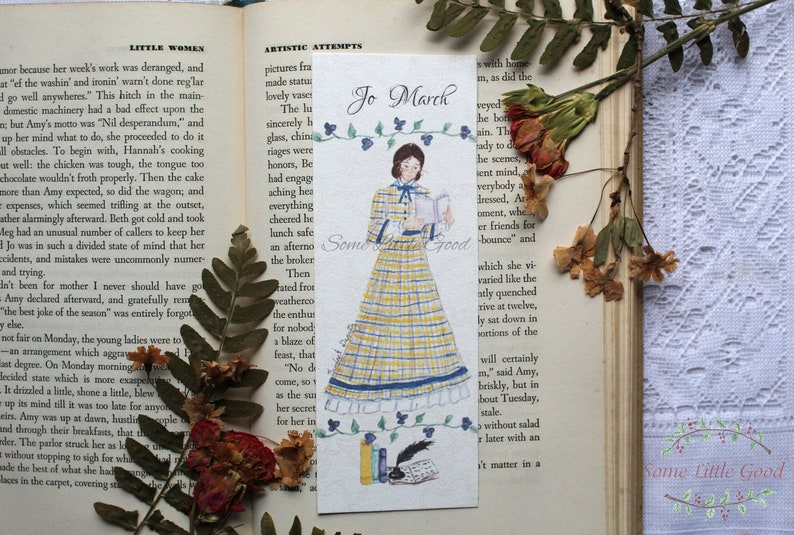 May include: A bookmark featuring a watercolor illustration of Jo March from Little Women. She is wearing a yellow and blue plaid dress and holding a book. The bookmark has the text "Jo March" at the top and "Some Little Good" at the bottom.