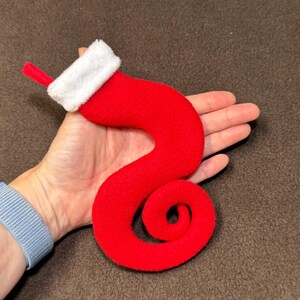 May include: A whimsical, red Christmas stocking with a white, fluffy cuff and a small red loop. The stocking is a unique, curved shape, perfect for holiday decor. The stocking is made of a soft, felt-like material.