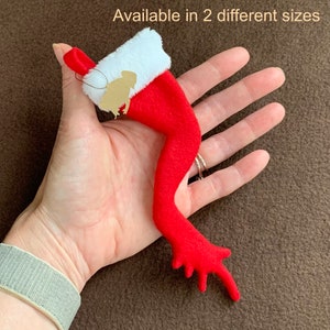 May include: Red felt stocking with a white cuff and a brown tag. The stocking is shaped like a leg and has a foot with toes. The stocking is held in a hand.