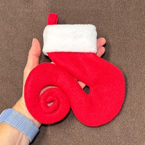 May include: A red felt Christmas stocking with a white fluffy cuff and a small loop for hanging. The stocking has a unique, swirled design. The background is a solid brown.