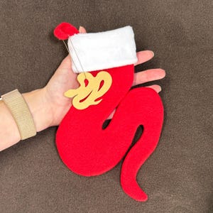 May include: A red Christmas stocking with a white cuff and a gold-colored decorative element. The stocking has a long, curved shape and a small red pom-pom at the top. A thin string is attached for hanging.