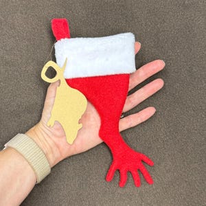 May include: A red and white Christmas stocking with a hand-shaped foot. The stocking is hanging from a string and has a gold paper tag attached to it.