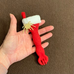 May include: A red felt Christmas stocking with a white cuff and a gold spider ornament. The stocking is shaped like an arm and hand. The spider is attached with a thin wire.