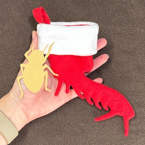 May include: A red felt Christmas stocking with a white cuff and a red loop. A gold-colored insect-shaped ornament is next to the stocking. The stocking has a long, stylized red shape extending from it.