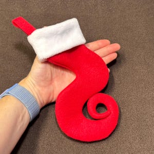 May include: A whimsical red Christmas stocking with a white cuff and loop for hanging. The stocking has a unique, swirled design. The stocking is made of a soft material, perfect for holiday decor.