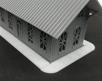 May include: A grey model building with a white pavement. The building has a grey roof and many windows. The building is on a black surface.