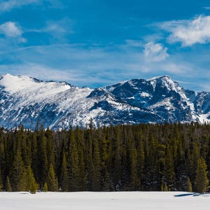 May include: A scenic panorama of snow-covered mountains against a vibrant blue sky dotted with clouds. A dense forest of evergreen trees forms the foreground, with a snowy expanse below. The image depicts a tranquil natural landscape.
