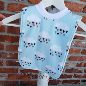 May include: A baby bib with a light blue background featuring white cloud designs with colorful heart-shaped raindrops. The bib has a white trim and is hanging on a white hanger. The text "Giggles of Love" is visible.
