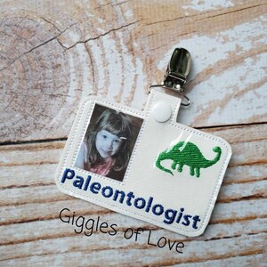 May include: White fabric badge with a photo of a child, a green dinosaur, and the text "paleontologist" in blue embroidery. The badge has a metal clip and the text "Giggles of Love" is printed below the badge.