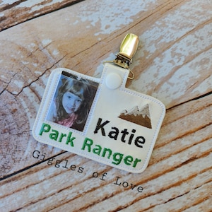 May include: White fabric badge with a photo of a young girl, the text "Park Ranger" in green, the name "Katie" in black, and a brown mountain graphic. The badge has a white button and a gold metal clip. The text "Giggles of Love" is printed below the badge.