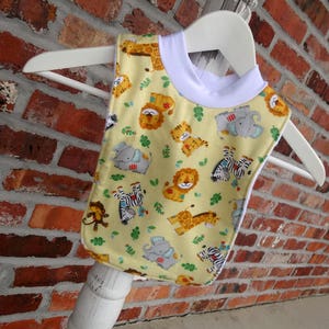 May include: A baby bib with a yellow background and a white trim. The bib is decorated with cartoon animals, including lions, zebras, elephants, and giraffes. The bib is hanging on a white hanger.