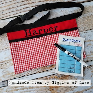May include: Red and white gingham fabric apron with a black strap and a red band with the name "Harper" embroidered on it. A blue and white guest check with a black dry erase marker is included. The text "Handmade Item by Giggles of Love" is at the bottom of the image.