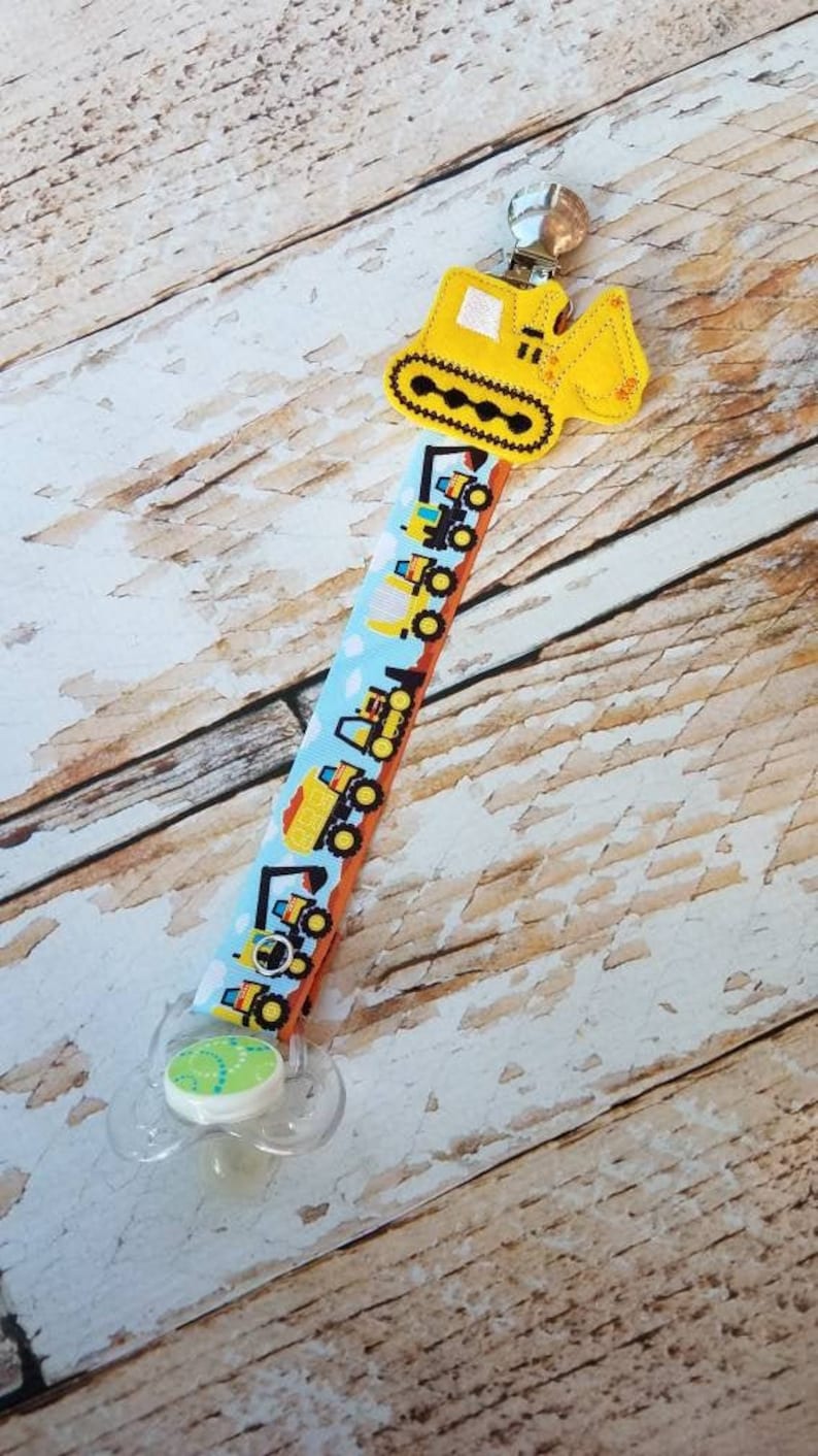 May include: A yellow construction vehicle pacifier clip with a blue and white ribbon featuring a repeating pattern of yellow construction vehicles. The clip is attached to a clear pacifier with a green and white design.