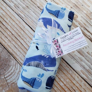 May include: A rolled-up baby receiving blanket with a whale pattern in shades of blue and white, tied with a white ribbon. The blanket has a tag that reads "Receiving Blanket" and "Giggles of Love."