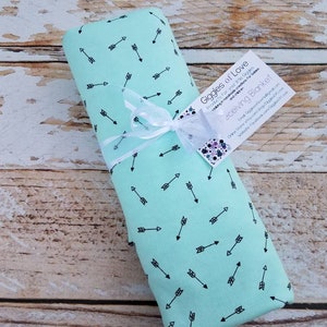 May include: A rolled-up mint green receiving blanket with a black arrow pattern, tied with a white ribbon. A tag reads "Giggles of Love" and "Receiving Blanket". The blanket is on a light brown wooden surface.