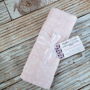 May include: A folded pink receiving blanket with a white floral pattern, tied with a white ribbon. A tag with the text "Giggles of Love" is attached. The blanket is on a wooden surface.
