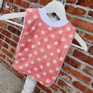 May include: A pink and white baby bib with a daisy print. The bib has a white trim around the neck.