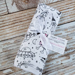 May include: A white receiving blanket with a black and white space-themed print. The print features rockets, stars, planets, and UFOs. The blanket is tied with a white ribbon and has a business card attached with the text "Giggles of Love" and the words "Receiving Blanket".