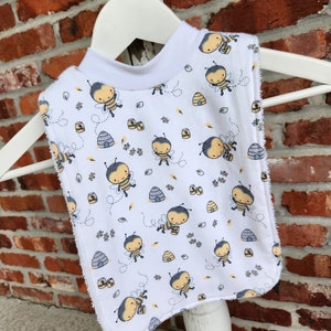 May include: White baby bib with a bee and honeycomb pattern. The bib has a ruffled edge and is made of soft fabric.
