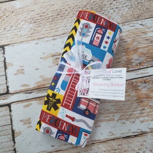 May include: A white receiving blanket with a colorful pattern of emergency vehicles, stars, and the words "Emergency!"  The blanket is folded and tied with a white ribbon. A small tag is attached to the ribbon with the text "Giggles of Love Products for your little Giggles. Specializing in handmade creations for babies and children. Receiving Blanket. Email: GigglesofLove@gmail.com. Online Store: etsy.com/shop/GigglesOfLove. Website: facebook.com/GigglesOfLove."