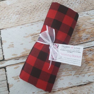 May include: A folded red and black buffalo plaid receiving blanket tied with a white ribbon. A small tag with the text "Giggles of Love" and "Receiving Blanket" is attached to the ribbon. The blanket is on a light wood surface.