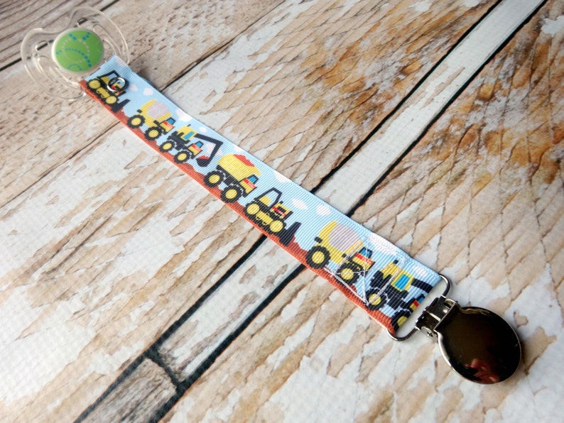 May include: A pacifier clip with a blue and white ribbon featuring a repeating pattern of yellow construction vehicles. The clip is attached to a silver pacifier clip.
