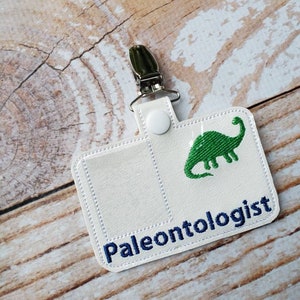 May include: White fabric badge holder with a green dinosaur and the text "Paleontologist" in blue embroidery. The badge holder has a metal clip and a white button.