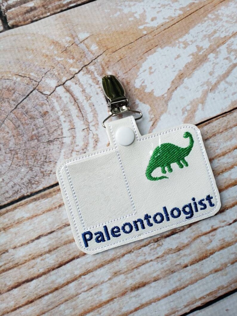 May include: White fabric badge holder with a green dinosaur and the text "Paleontologist" in blue embroidery. The badge holder has a metal clip and a white button.