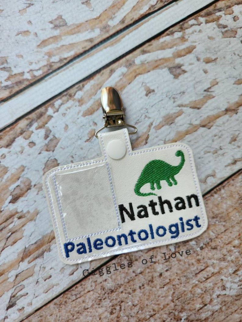 May include: White fabric badge holder with a clear plastic window, a green dinosaur graphic, and the text "Nathan Paleontologist". The badge holder has a metal clip on the top and is made by Giggles of Love.