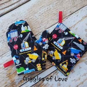 May include: A quilted oven mitt and pot holder set. The black fabric is patterned with construction vehicles in yellow, blue, and red. The set has red hanging loops and the text "Giggles of Love".