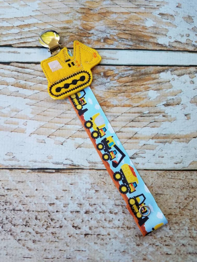 May include: Yellow felt construction vehicle clip with a blue and white ribbon featuring a repeating pattern of construction vehicles.