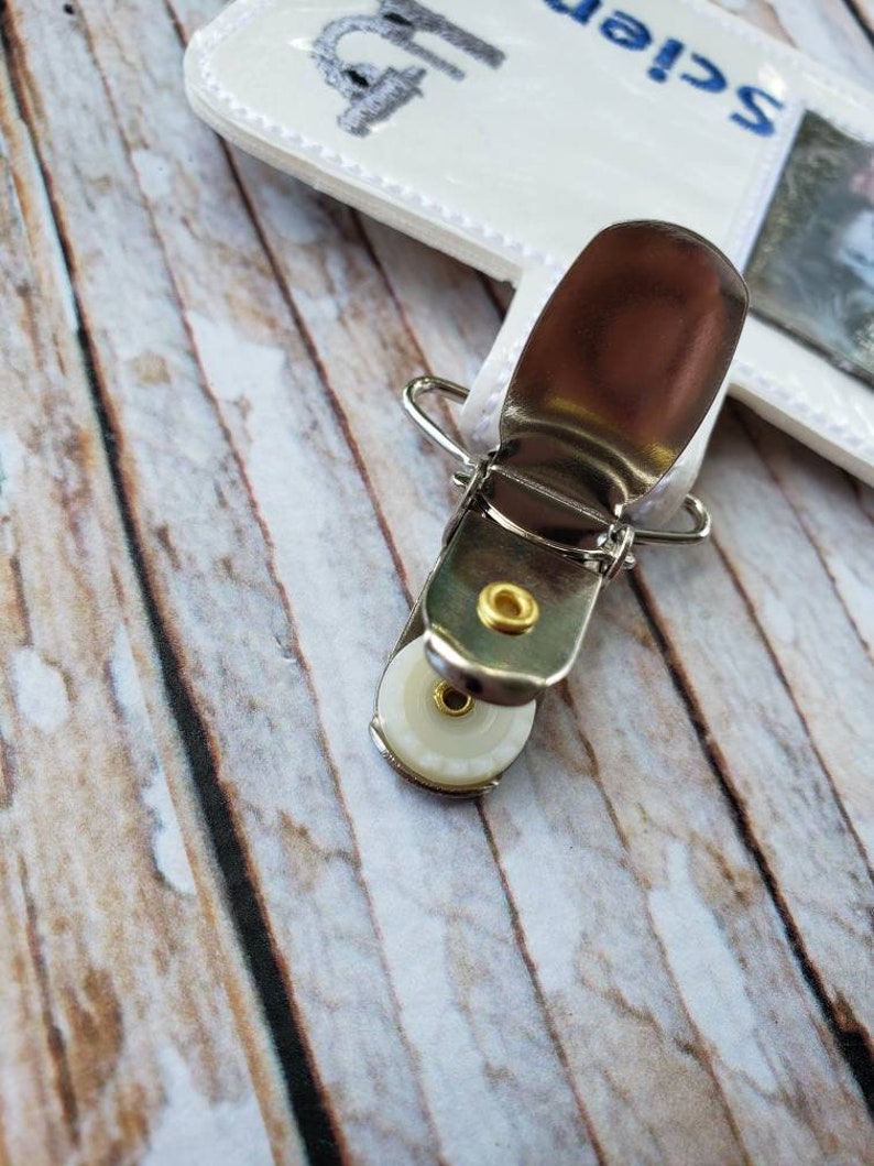 May include: A silver metal clip with a white plastic wheel. The clip is attached to a white fabric strap with the word "Science" embroidered on it.