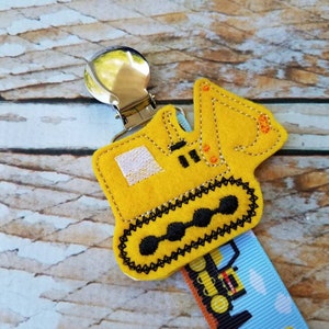 May include: Yellow felt construction vehicle clip with a silver clip. The vehicle is a bulldozer with black tracks and orange details. The clip is attached to a blue and white ribbon with a repeating pattern of construction vehicles.