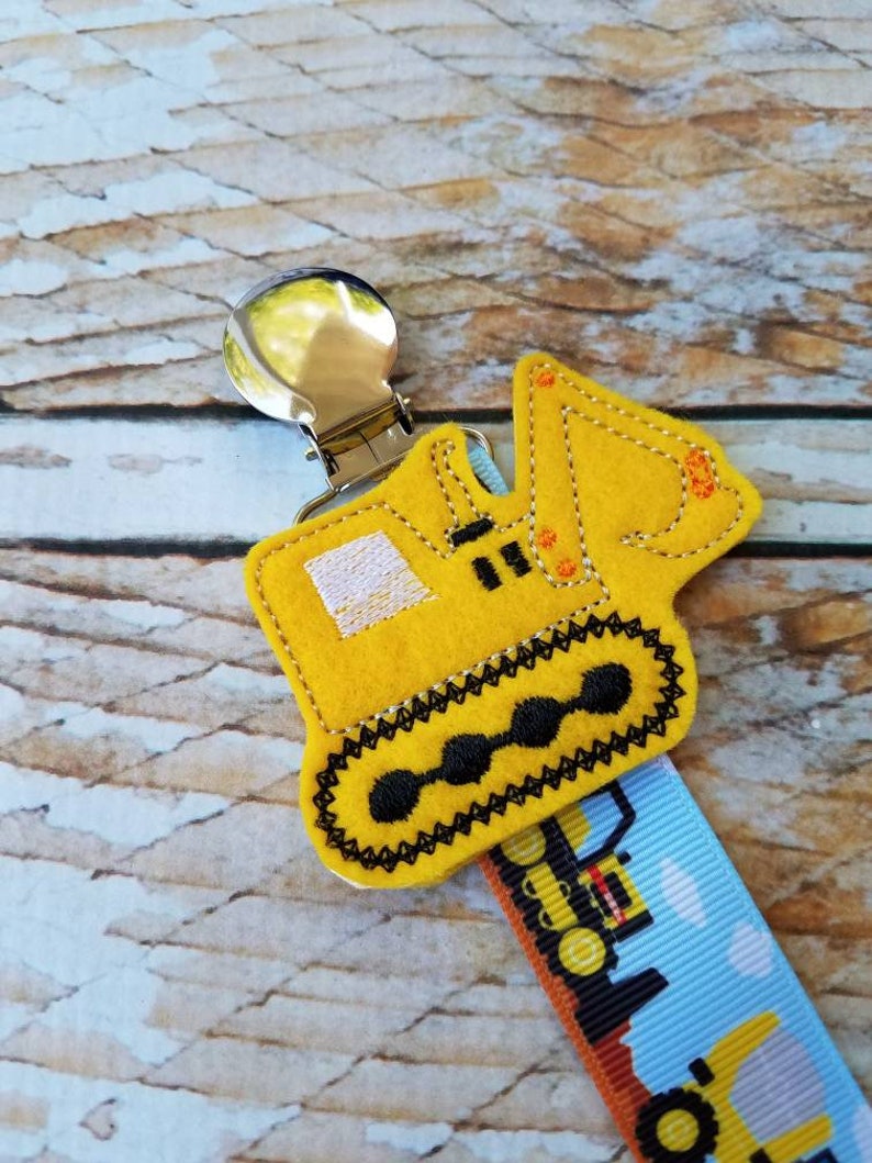 May include: Yellow felt construction vehicle clip with a silver clip. The vehicle is a bulldozer with black tracks and orange details. The clip is attached to a blue and white ribbon with a repeating pattern of construction vehicles.