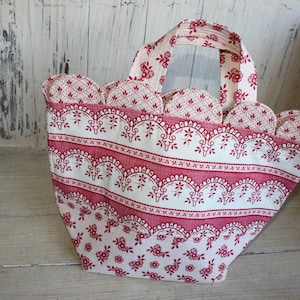Peut inclure: Un sac cabas en tissu rouge et blanc avec un bord festonné et un motif floral. Le sac a une anse et est fait d'un matériau léger.