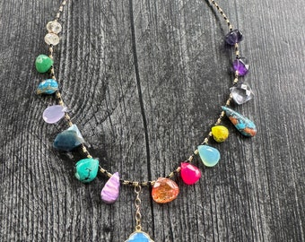 Puede incluir: Un collar de piedras preciosas de colores con una variedad de piedras, incluyendo turquesa, amatista y ágata. El collar está hecho con cuentas negras y una cadena dorada. El collar tiene un gran colgante de piedra azul claro.