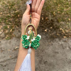 May include: A green and gold car charm featuring a religious image of a saint with a white tassel. The charm is decorated with green and gold flowers.