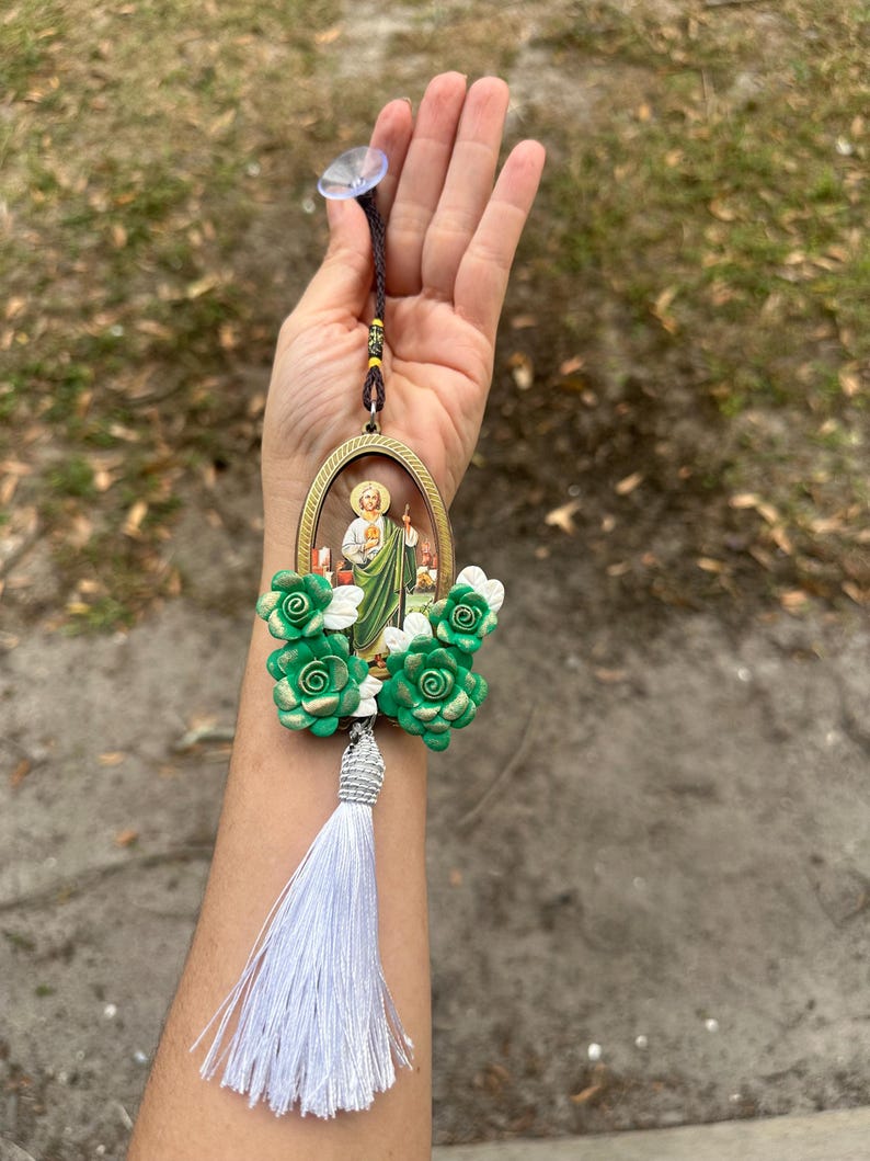 May include: A green and gold car charm featuring a religious image of a saint with a white tassel. The charm is decorated with green and gold flowers.