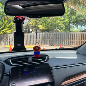 May include: The interior of a car with a black dashboard and a screen displaying "iPhone (2)" and "NW 07:11:01 70". A small, colorful, crocheted bucket hangs from the rearview mirror.