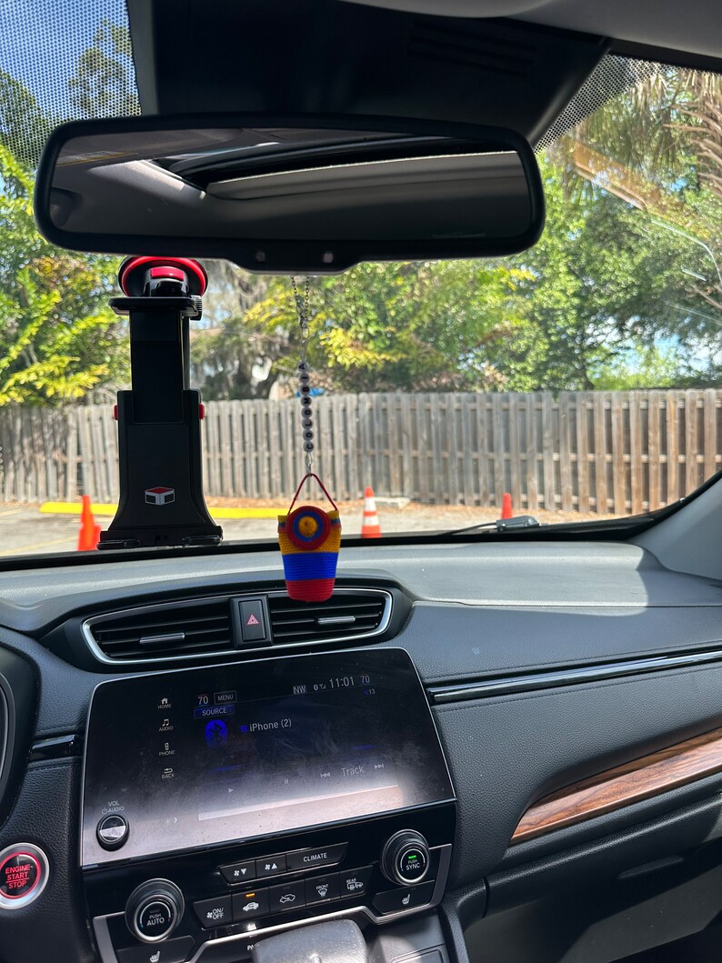May include: The interior of a car with a black dashboard and a screen displaying "iPhone (2)" and "NW 07:11:01 70". A small, colorful, crocheted bucket hangs from the rearview mirror.