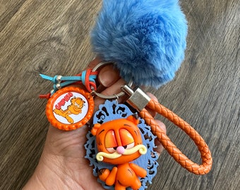 May include: A blue fuzzy keychain with a Garfield character charm. The charm is orange and has a blue frame. The keychain also has a small orange and blue charm with the word "Garfield" on it.