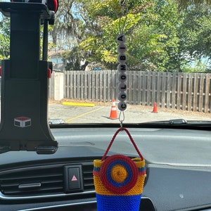 May include: A rearview mirror car charm with a woven bag in the colors of the Colombian flag, red, yellow, and blue, with a yellow circle in the center. The charm is hanging from a chain with white beads that spell out "COLOMBIA".