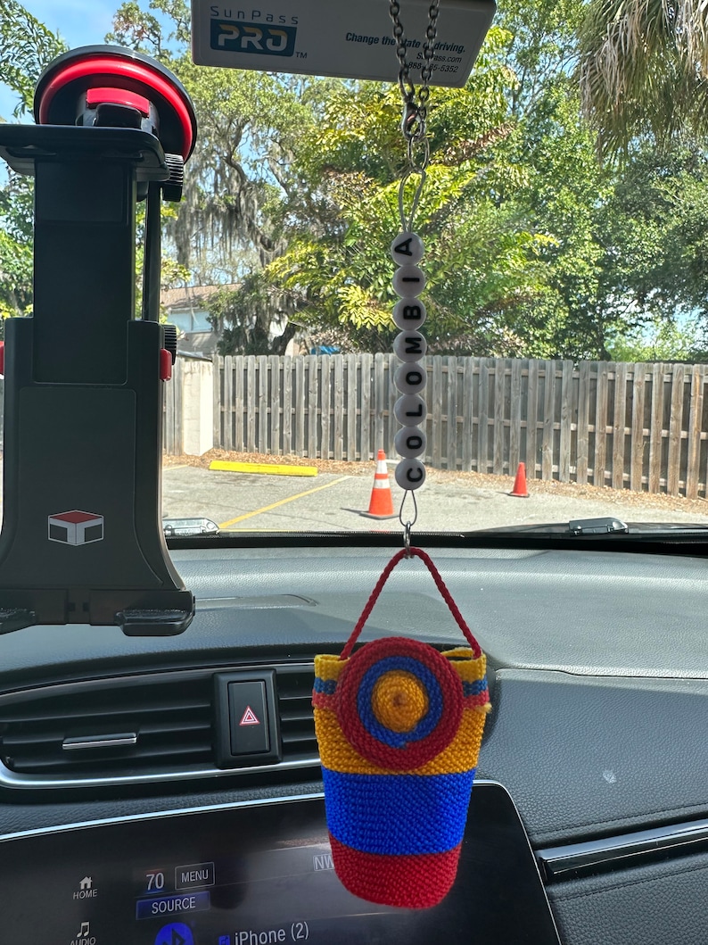 May include: A rearview mirror car charm with a woven bag in the colors of the Colombian flag, red, yellow, and blue, with a yellow circle in the center. The charm is hanging from a chain with white beads that spell out "COLOMBIA".