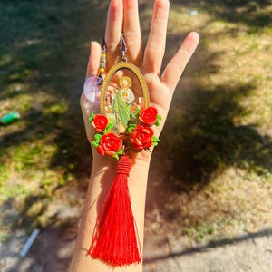 May include: A red tassel hangs from a gold oval frame with a religious image inside. The frame is surrounded by red plastic flowers.