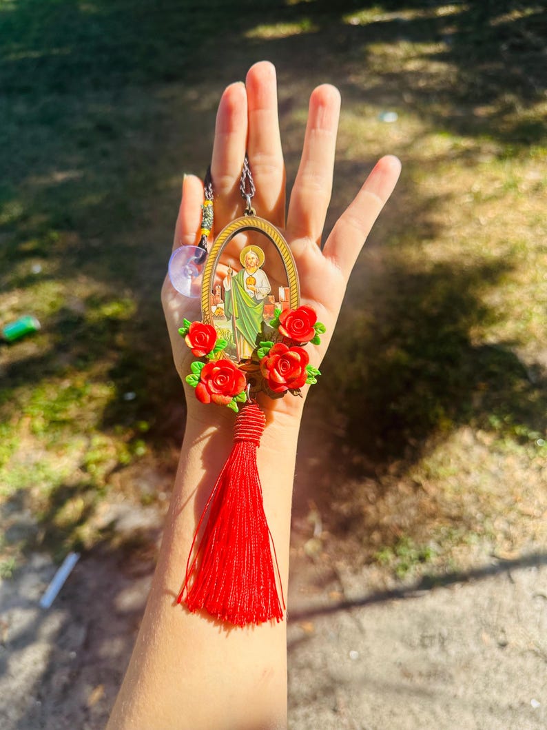 May include: A red tassel hangs from a gold oval frame with a religious image inside. The frame is surrounded by red plastic flowers.
